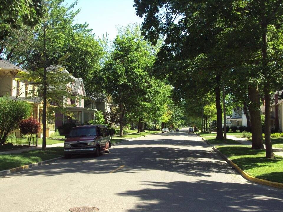 A residential street of older two-storey houses with mature trees and front lawns.
